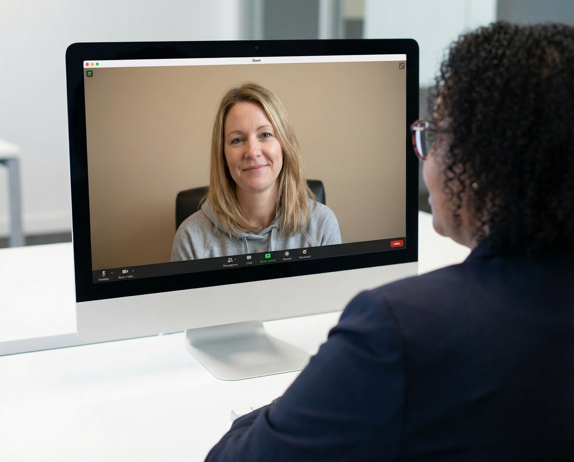 Woman in a dark blazer at a desk, looking at a computer screen showing another woman in a video call.