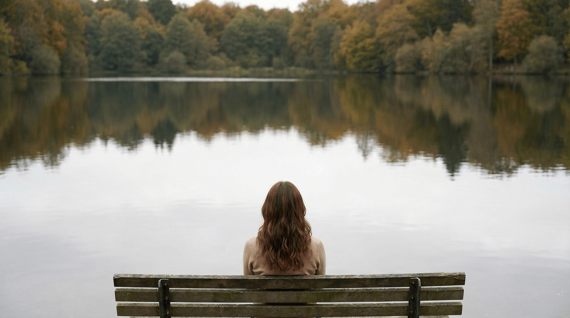 Woman with long brown hair sits on a bench, looking at a calm lake surrounded by fall trees.