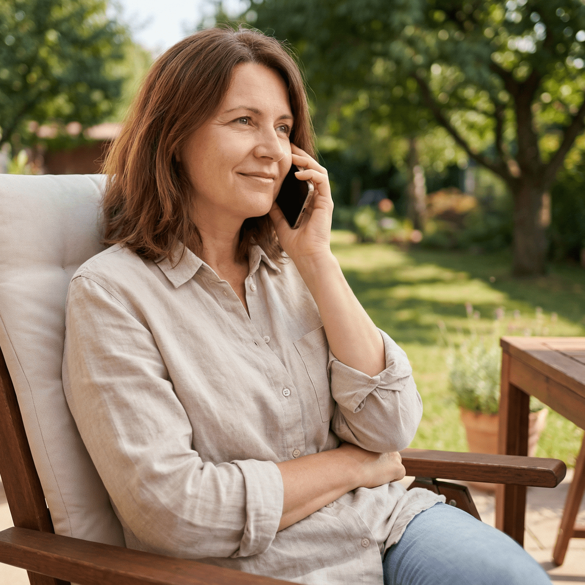 Woman in a chair in a garden, holding a phone to her ear and smiling.