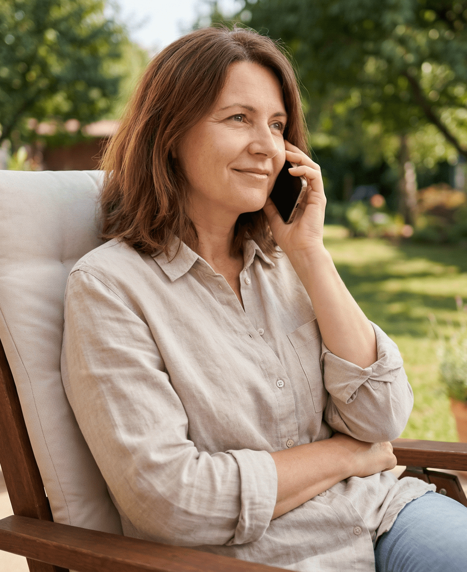 Woman in a chair in a garden, holding a phone to her ear and smiling.
