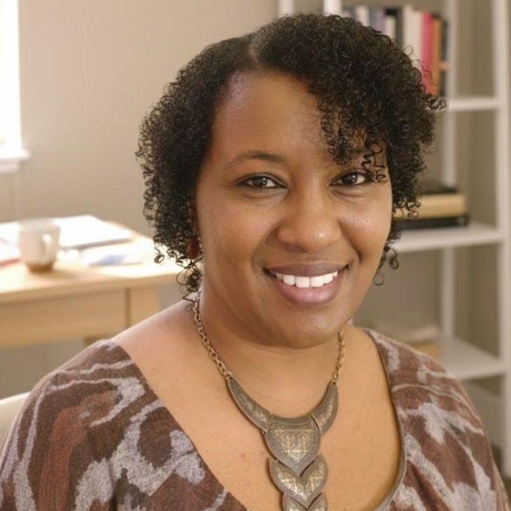 Woman smiling indoors, wearing a necklace and patterned top, with curly hair and a neutral background.