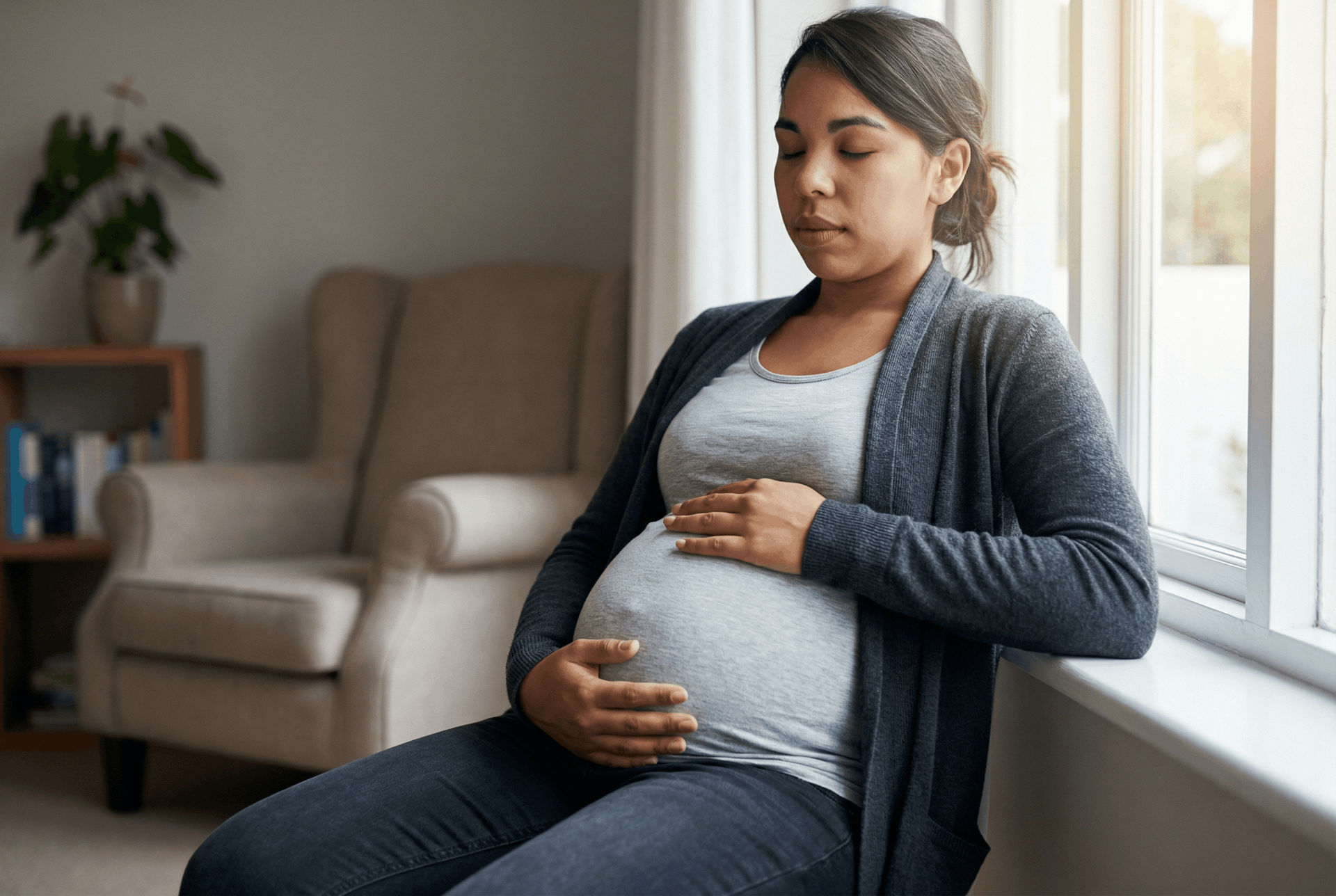 Pregnant person sitting by a window, hands on her abdomen, serene expression.