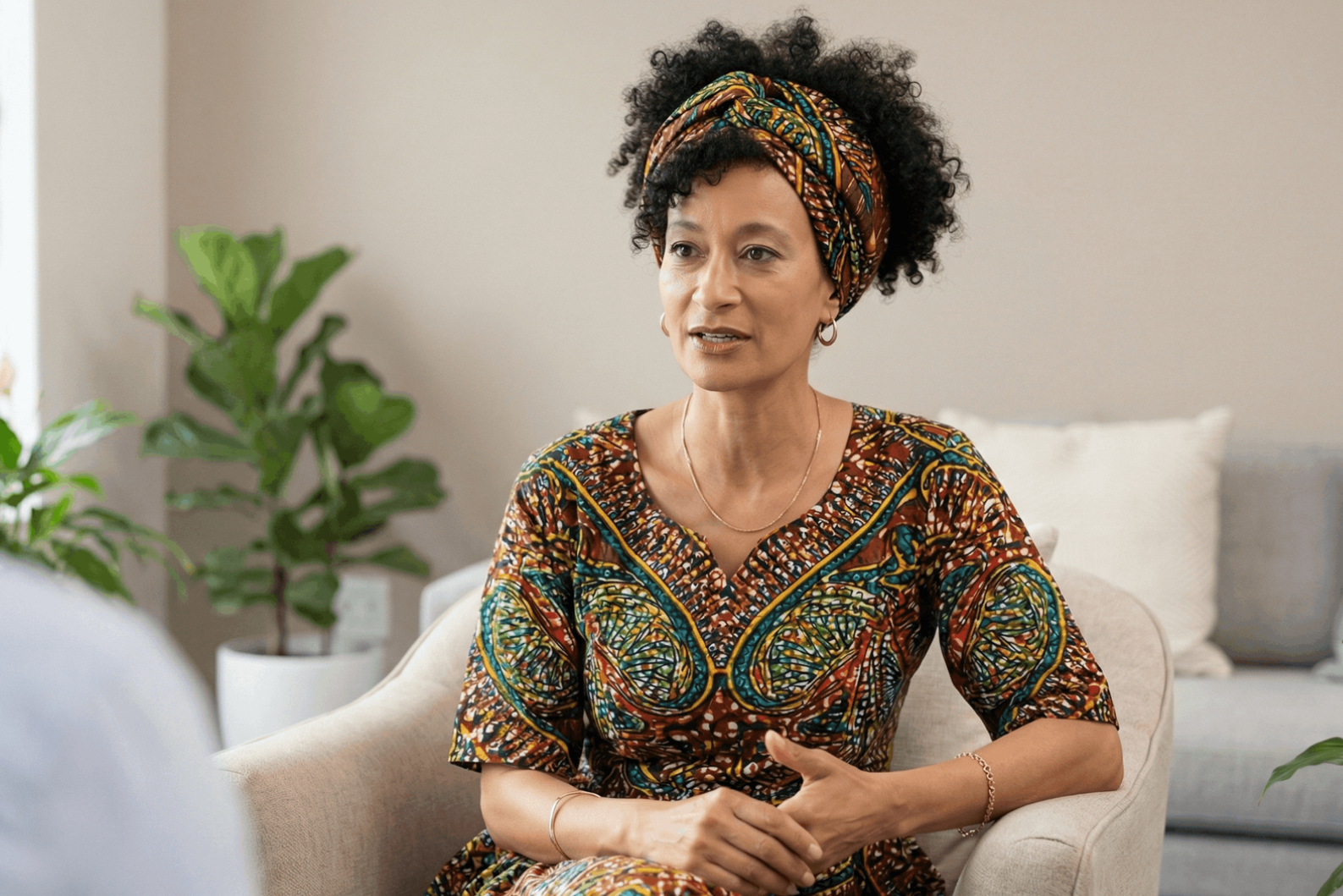 Woman in patterned dress and headscarf speaks while seated in a chair.