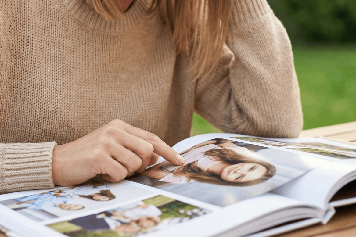 Woman in beige sweater looks at photo album outdoors, pointing at a picture.