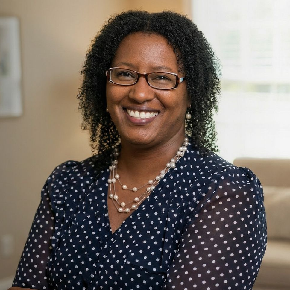Woman with glasses smiles, wearing polka dot blouse and pearl necklace.