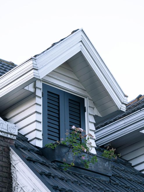 A white gable dormer window with dark blue louvered shutters and a flower box on a dark tiled roof.