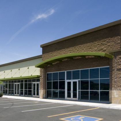 A strip mall storefront with tan stucco walls, green metal awnings, large glass windows, and a handicap parking spot.