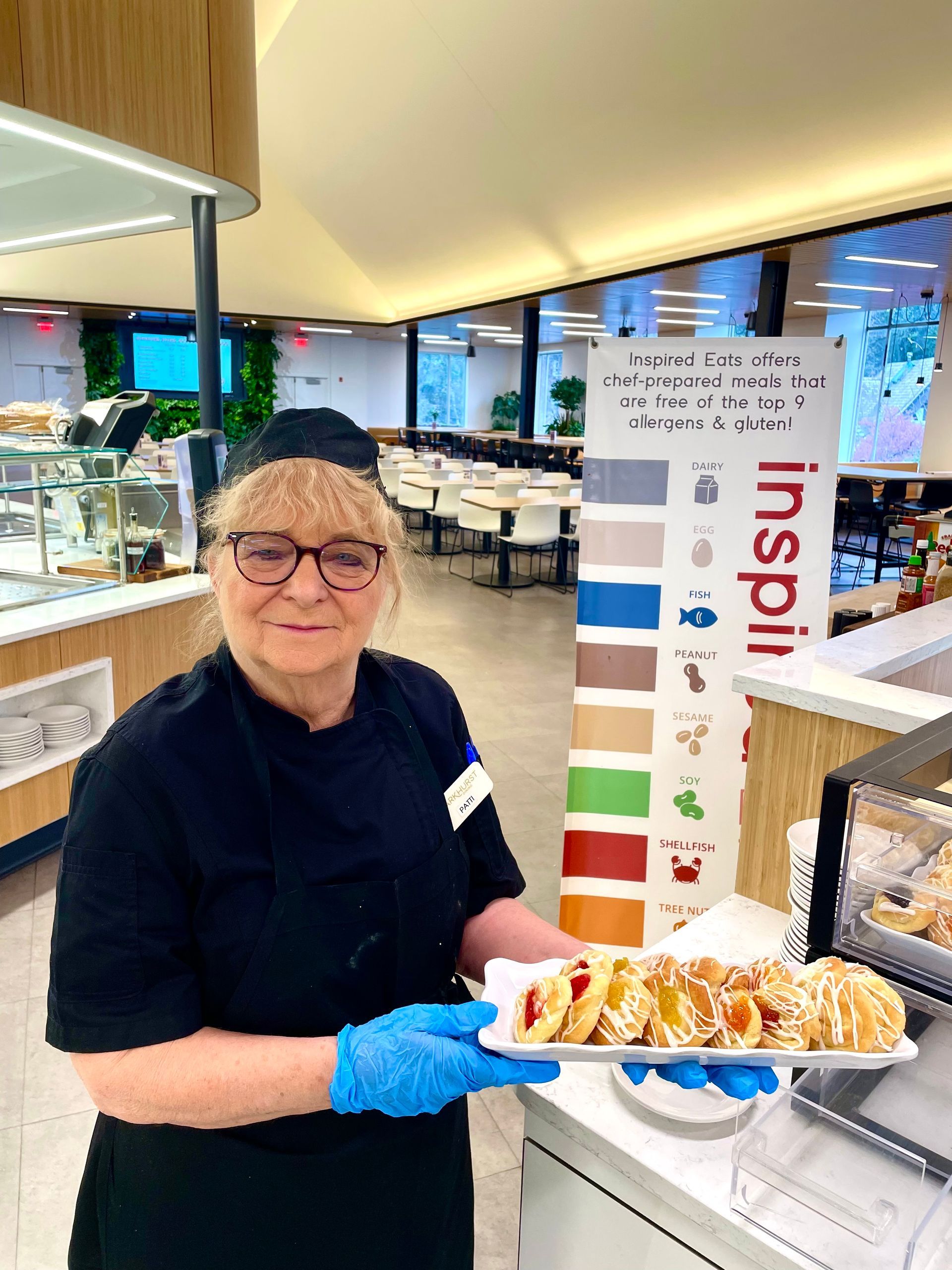 Woman in uniform holding tray of pastries in a brightly lit cafe with allergen Inspired eats sign.
