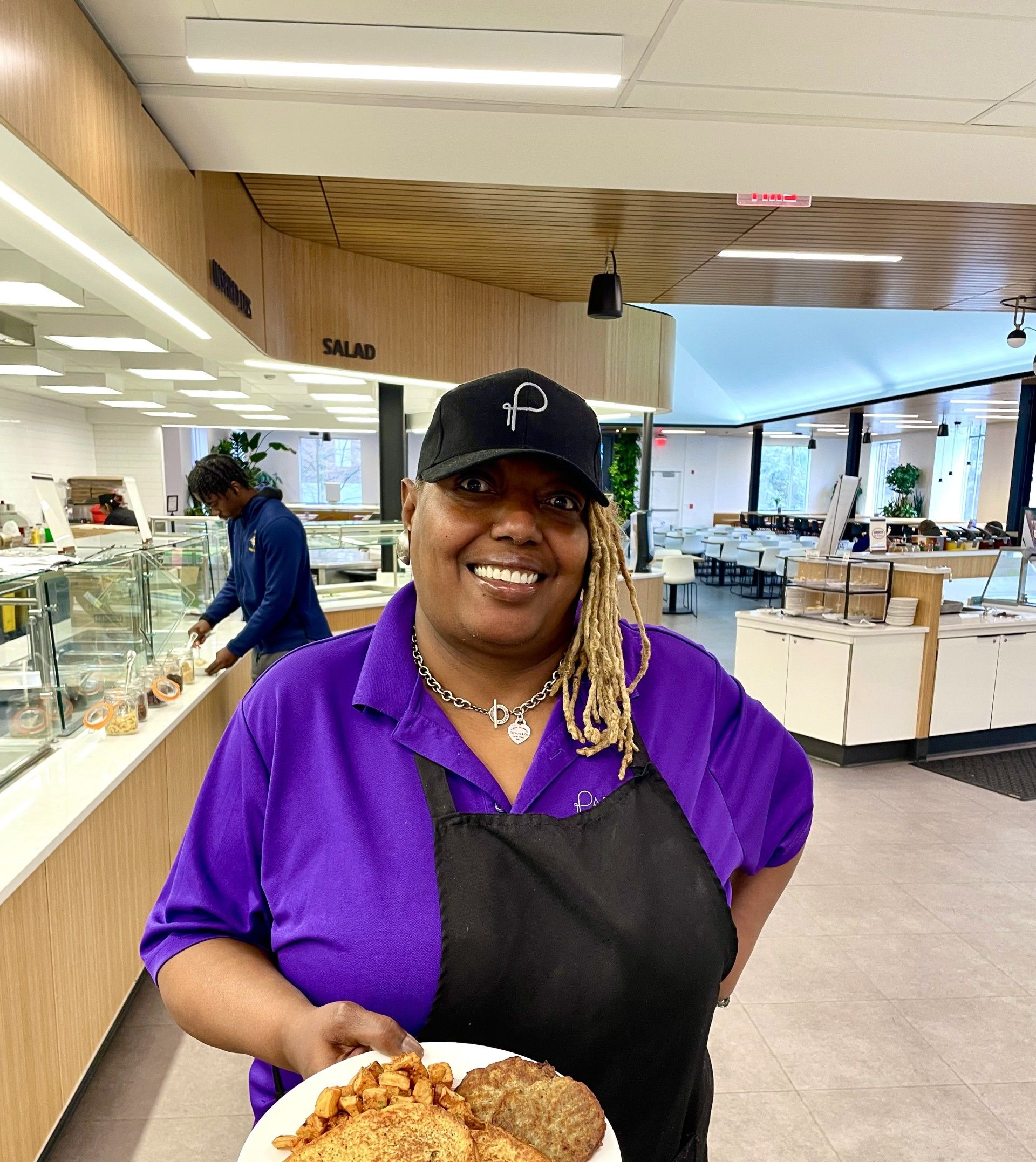 Woman in purple shirt, black apron, holding plate of food, smiling in a modern cafeteria.