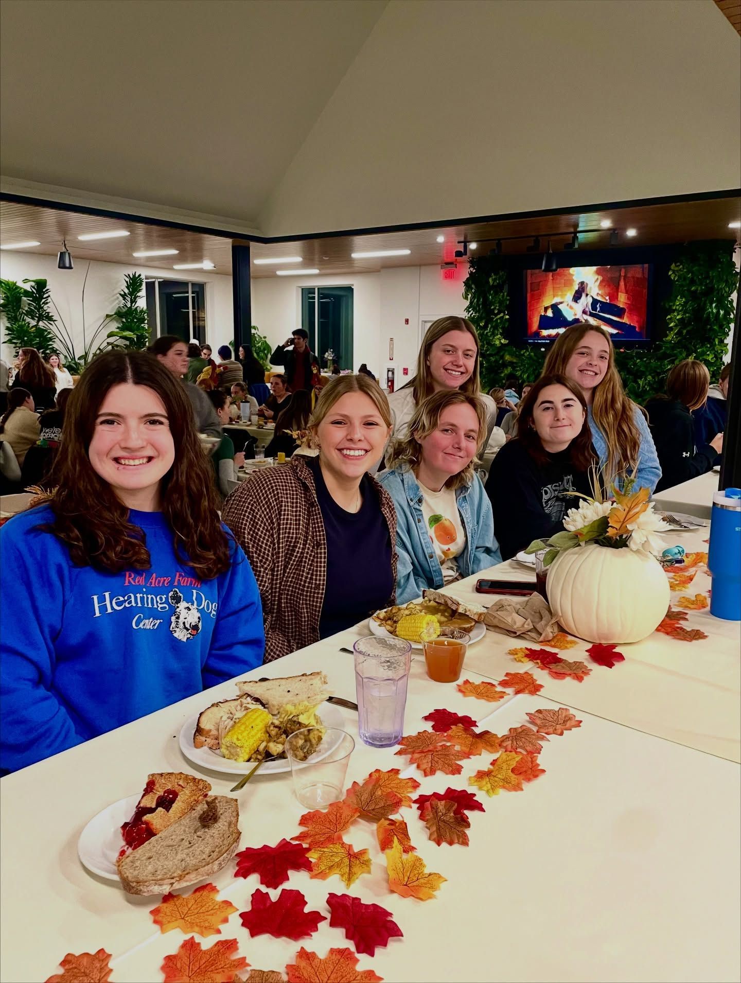 Six people sit at a festive table decorated with artificial autumn leaves, food, and a white pumpkin in a dining hall.