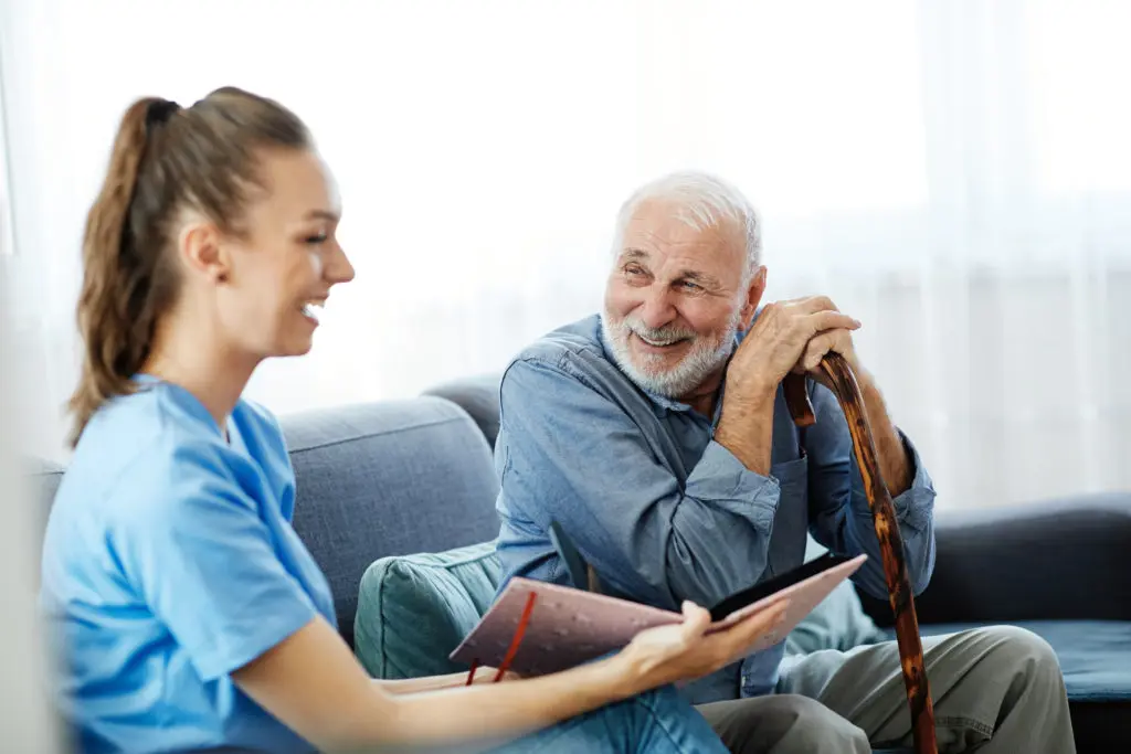 A nurse is sitting on a couch talking to an elderly man with a cane.