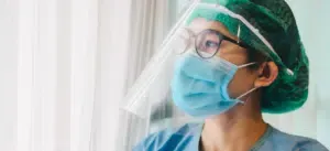 A nurse wearing a mask and a face shield is looking out of a window.