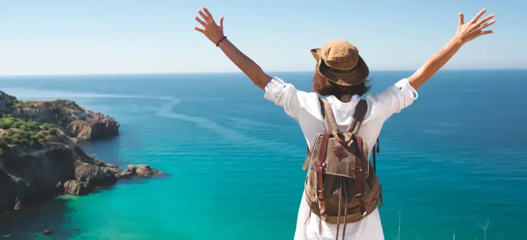 A woman with a backpack is standing on top of a hill overlooking the ocean with her arms outstretched.
