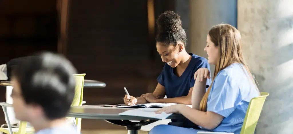 Two nurses are sitting at a table looking at a tablet.