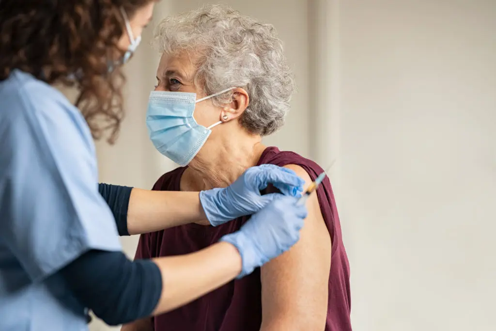 An elderly woman wearing a mask is getting a vaccine from a nurse.