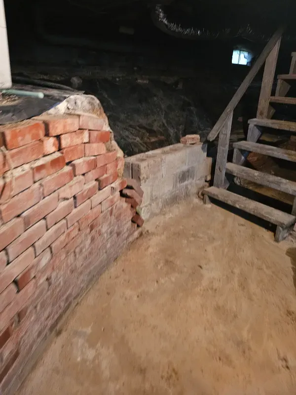 Brick wall damage near a wooden staircase in a dimly lit basement.