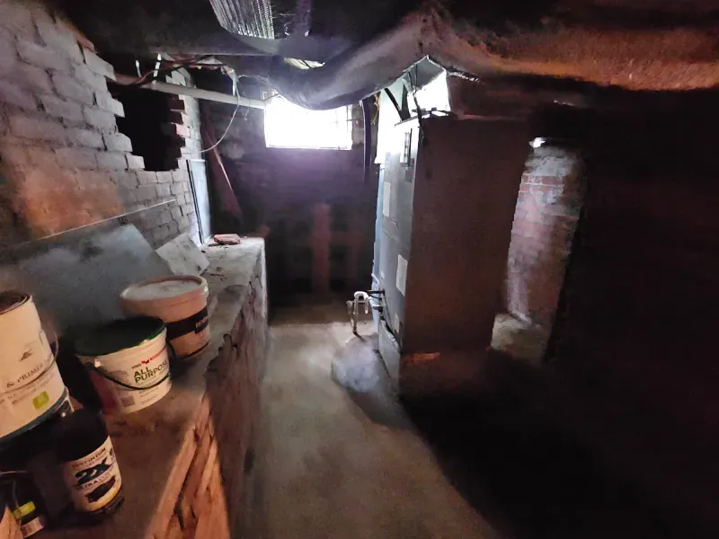 Basement hallway with brick walls, old furnace, and paint cans. Sunlight streams in.
