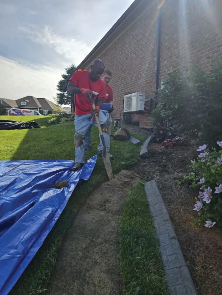 Two men digging a trench along a house's foundation, blue tarp in the foreground, sunny day.
