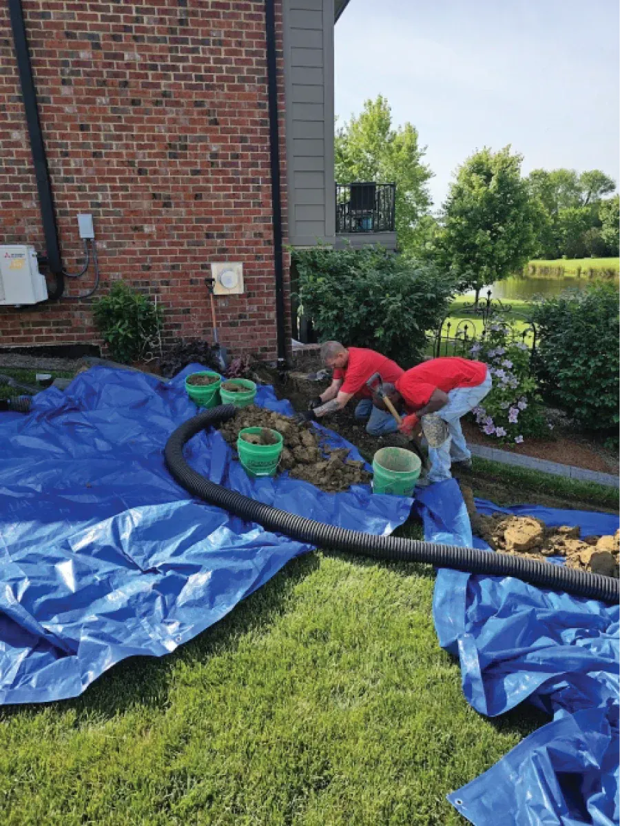 Two people in red shirts working on a landscaping project next to a brick building and lawn.