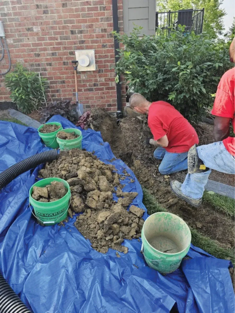 Men in red shirts installing drainage, digging a trench near a brick building.