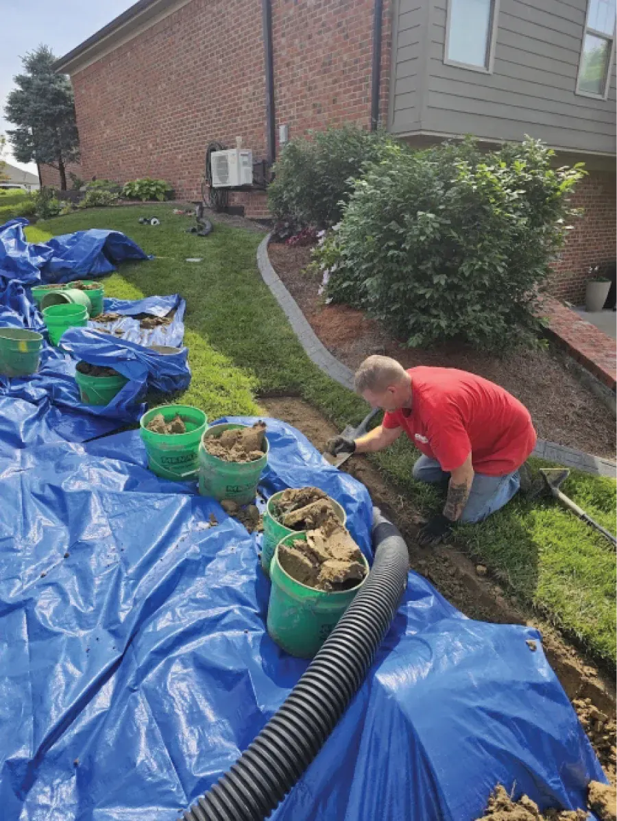Man kneeling, installing drainage pipe in yard near house. Blue tarp, buckets of soil, green grass.