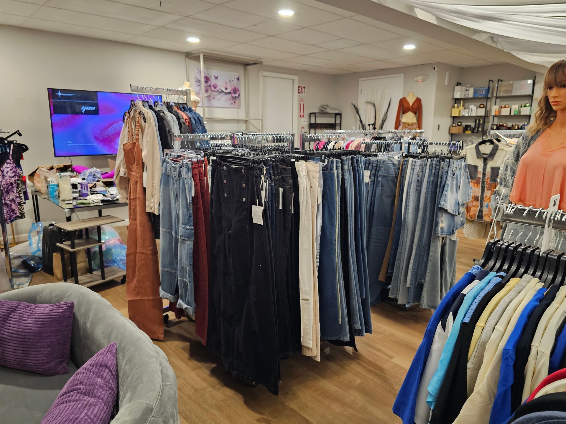 A clothing store interior featuring racks of denim pants, tops, and a mannequin near a purple couch and a TV display.