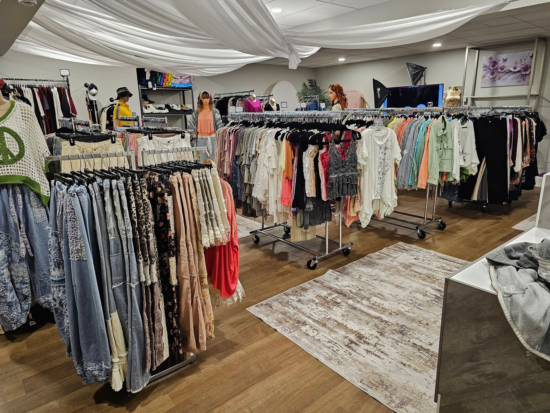 A retail clothing boutique interior featuring racks of organized apparel, a wooden floor, and draped white ceiling fabric.
