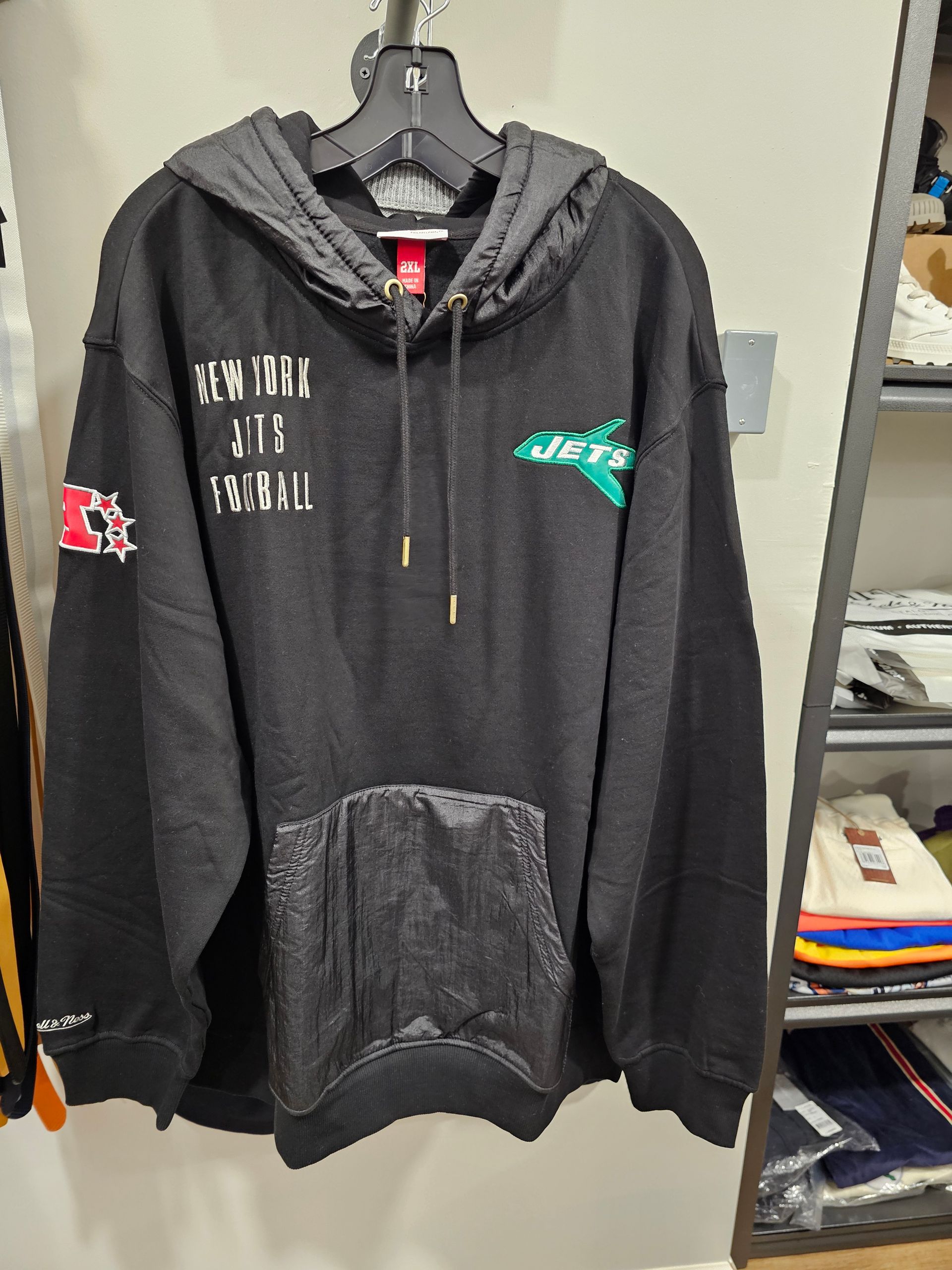 A black hooded sweatshirt with white text, a green logo on the chest, a red patch on the sleeve, and a textured pocket.