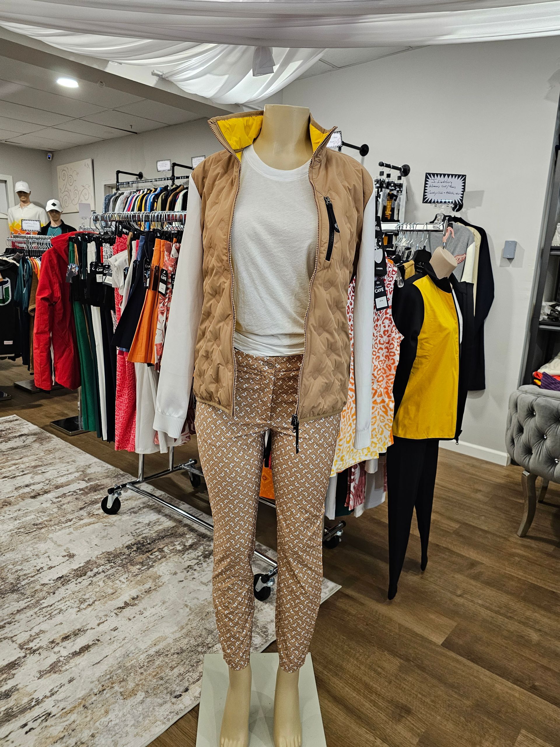 A mannequin wears a white long-sleeve top, a tan quilted vest with a yellow collar, and matching patterned tan joggers.