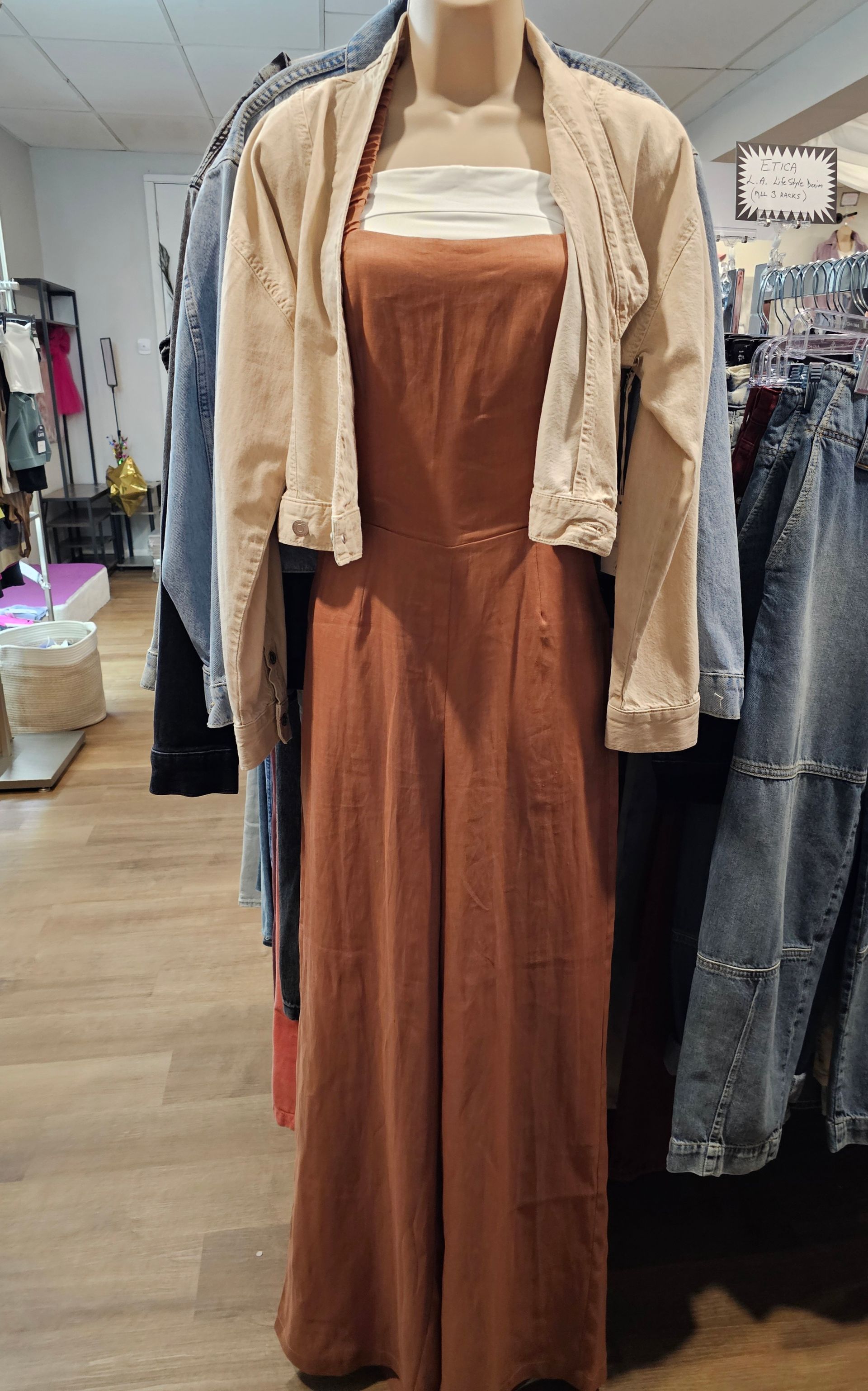 A mannequin in a clothing store wears a tan cropped jacket over a rust-colored, wide-leg jumpsuit with a white top.