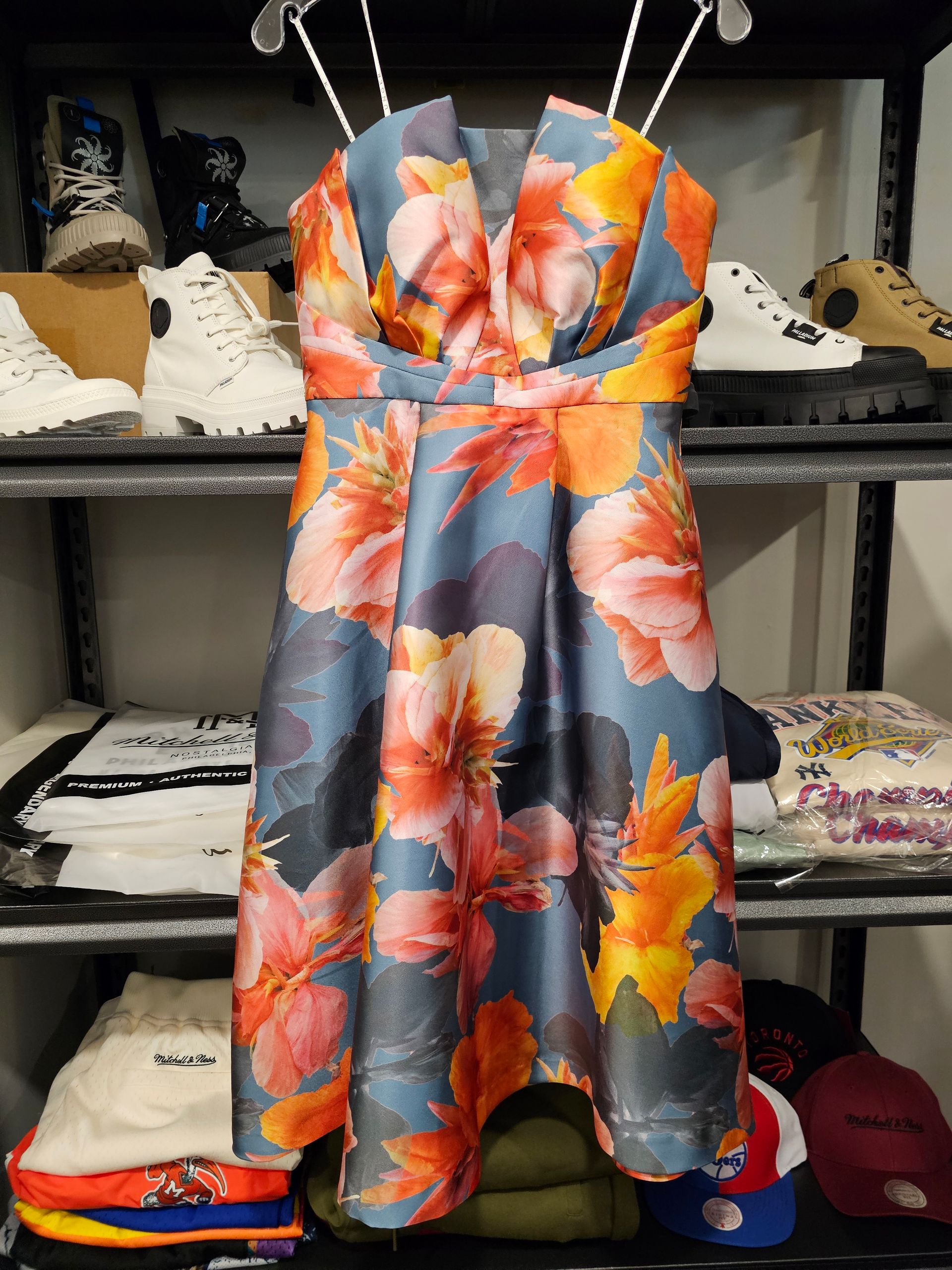 A strapless, tea-length floral dress with orange, pink, and yellow petals on a slate blue background, displayed on a rack.
