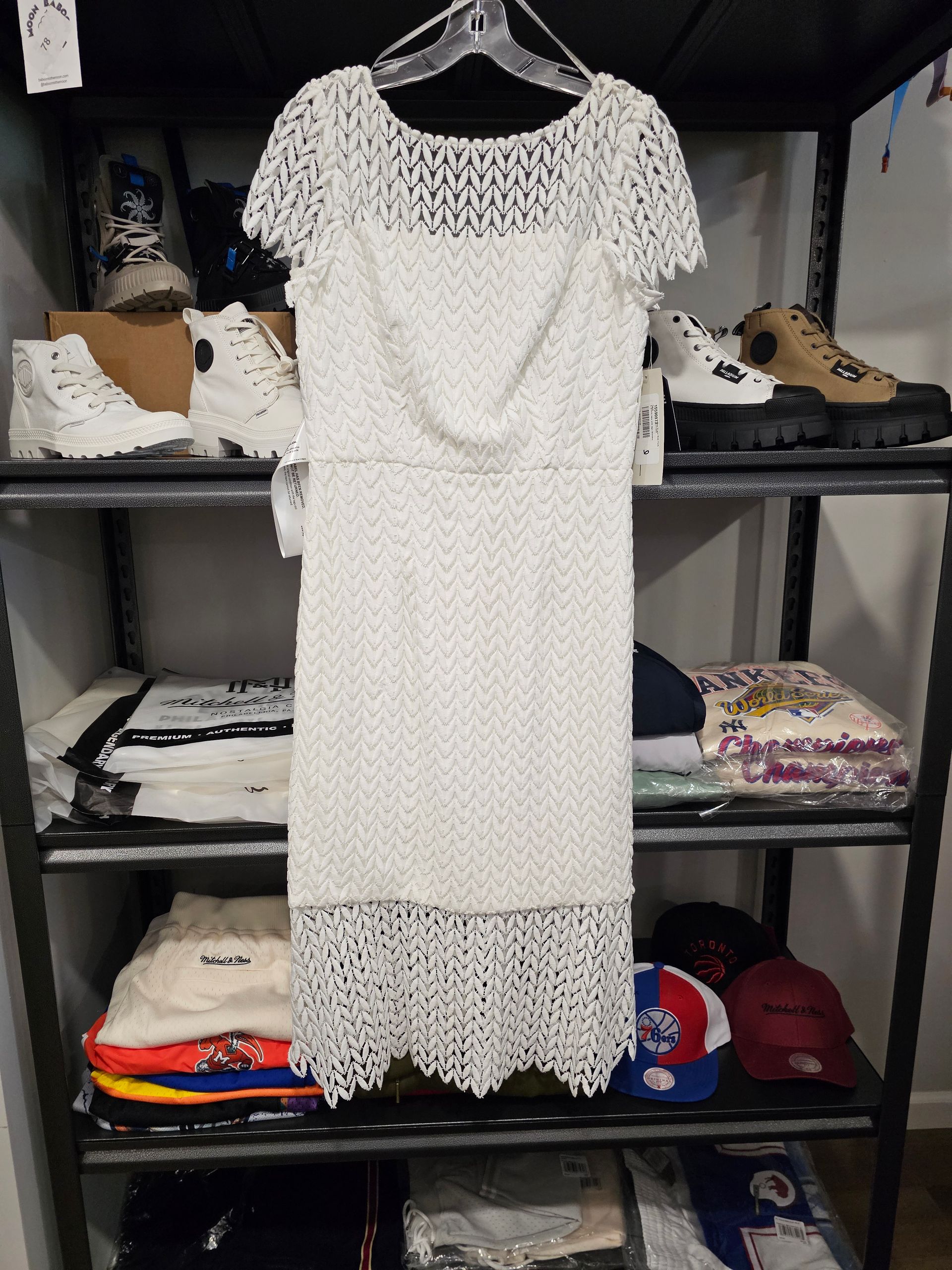 A white, knee-length, crochet-style dress with short sleeves hanging on a metal shelf unit filled with clothing and shoes.