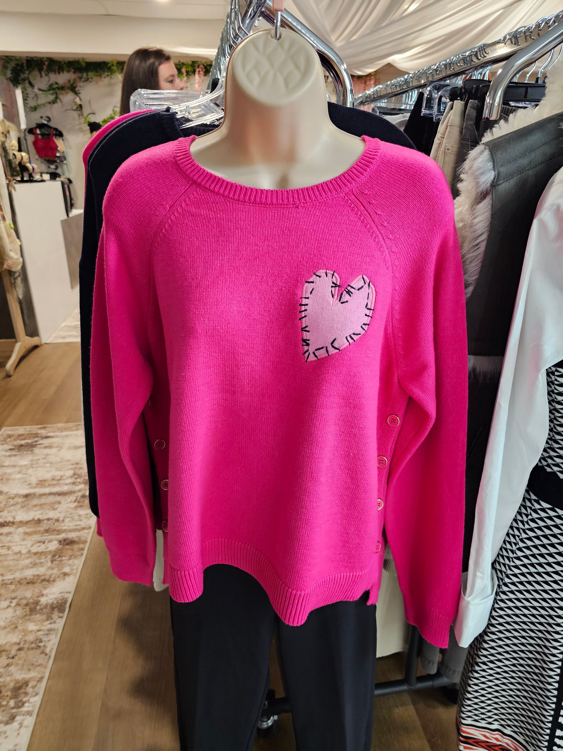A vibrant pink long-sleeved sweater with a textured heart patch on a display mannequin in a clothing store.