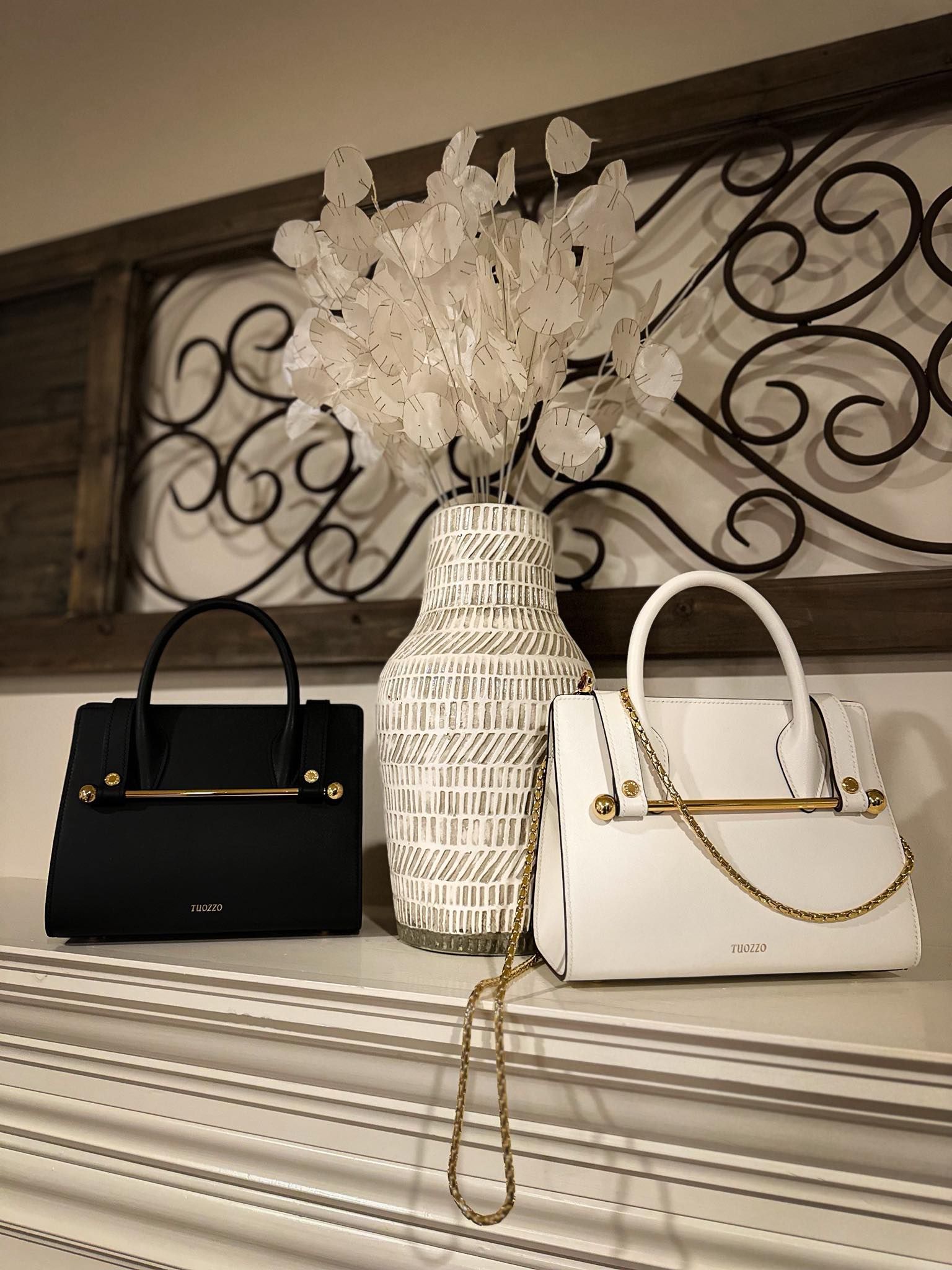 Black and white handbags on a shelf, with a vase of white flowers and decorative metal scrollwork in the background.