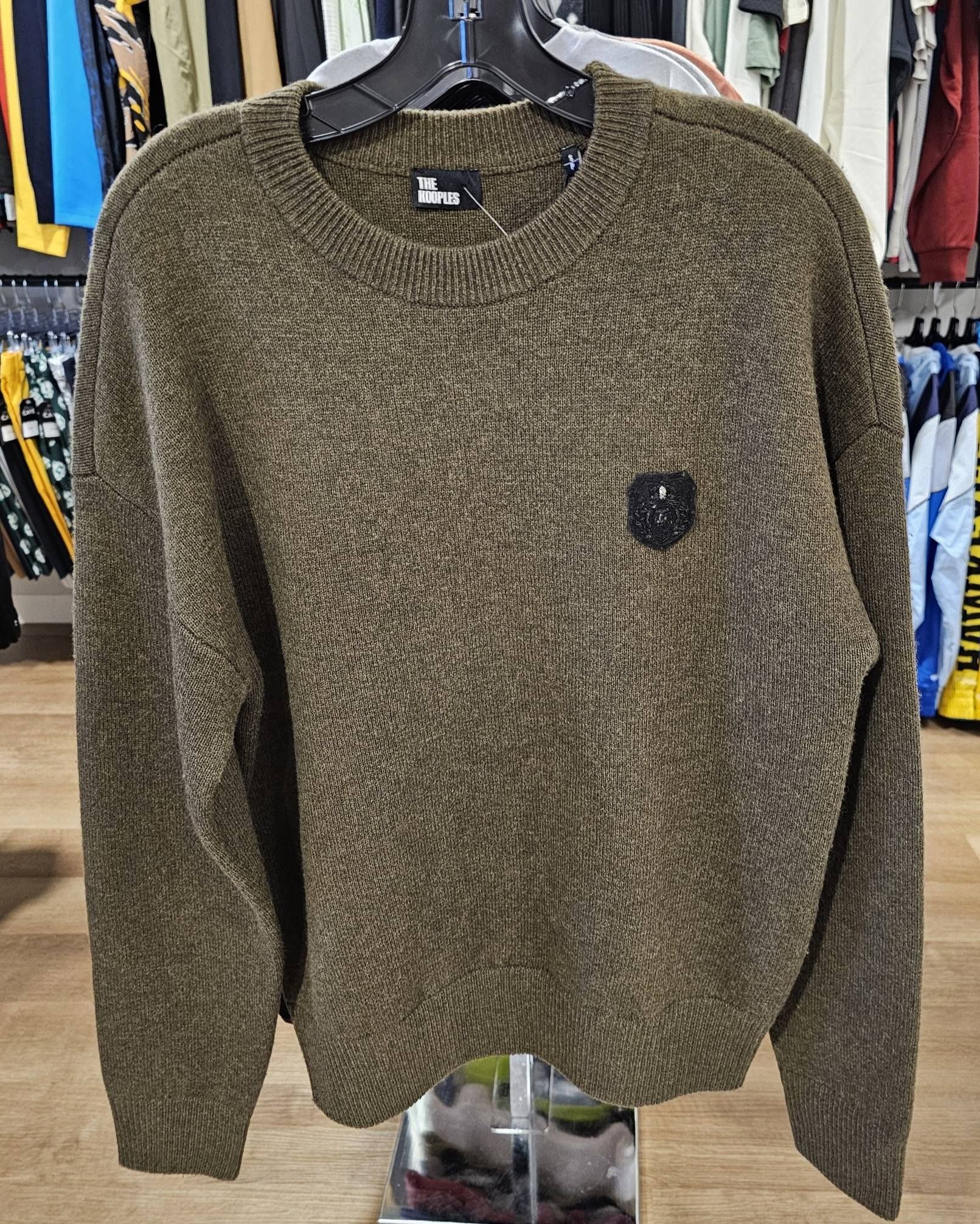 Olive-green textured sweater on a hanger, small emblem on the chest. Clothing store setting.