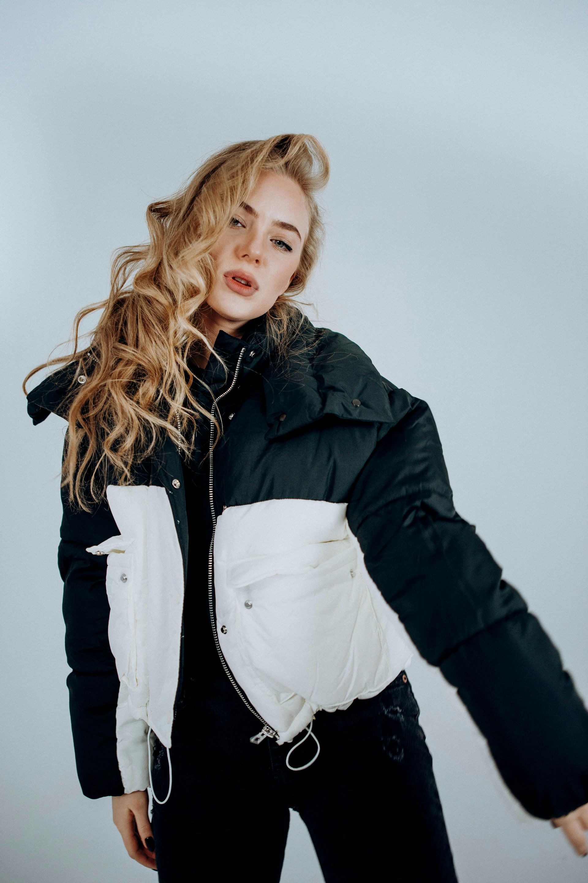 Blond woman in black and white puffer jacket, curly hair, posing against a light gray background.