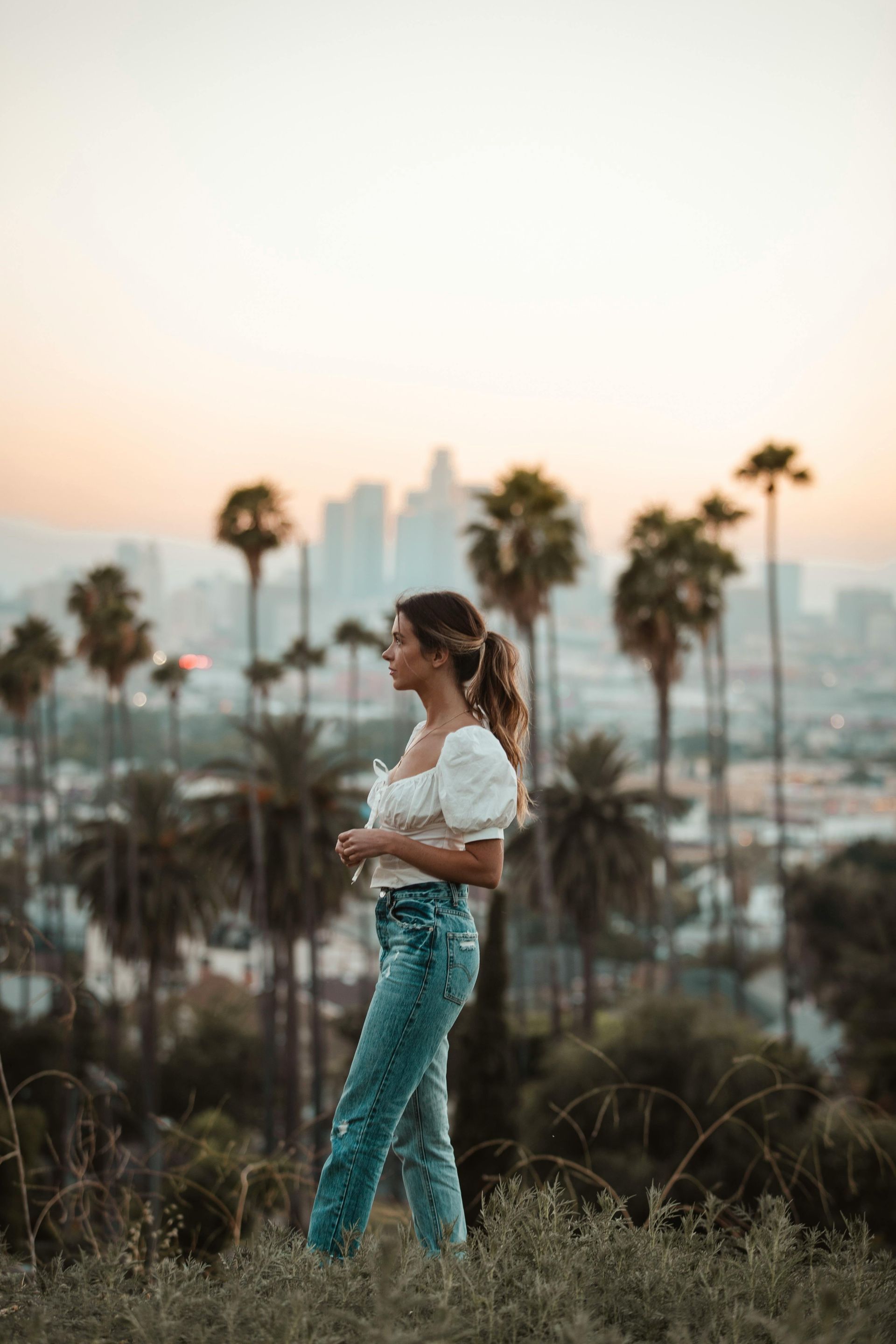Woman in white top and jeans poses outdoors, city skyline in the background at sunset.