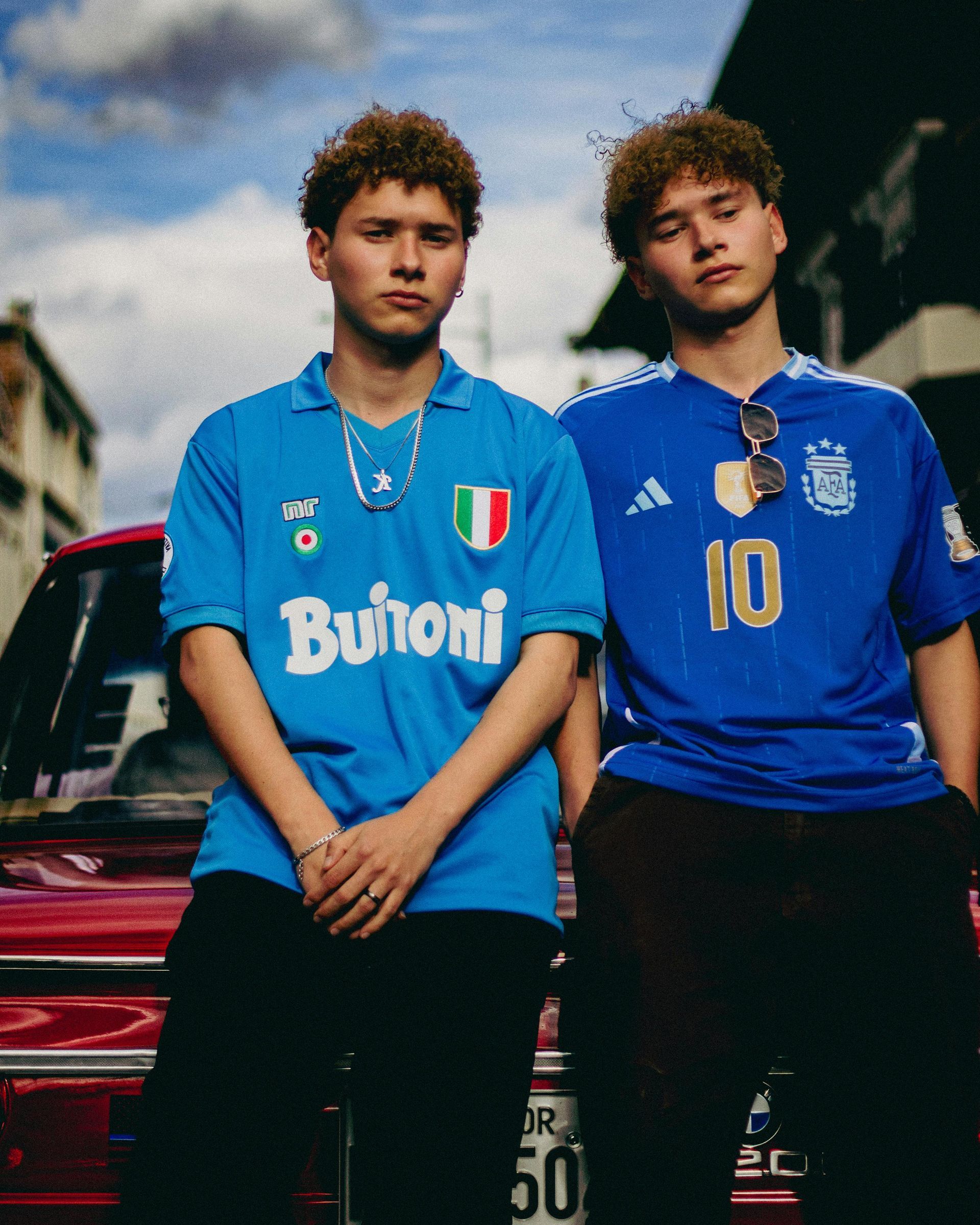 Two young men wearing soccer jerseys, posing next to a red car, outdoor setting.