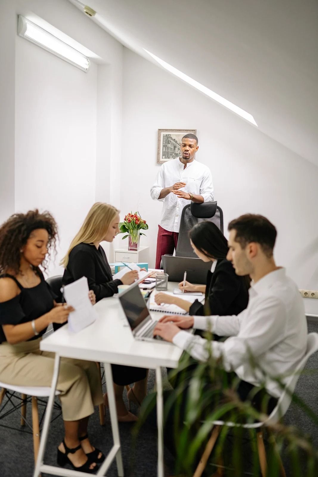 Four people in a bright office meeting; one person stands presenting while three sit at a white table using laptops.