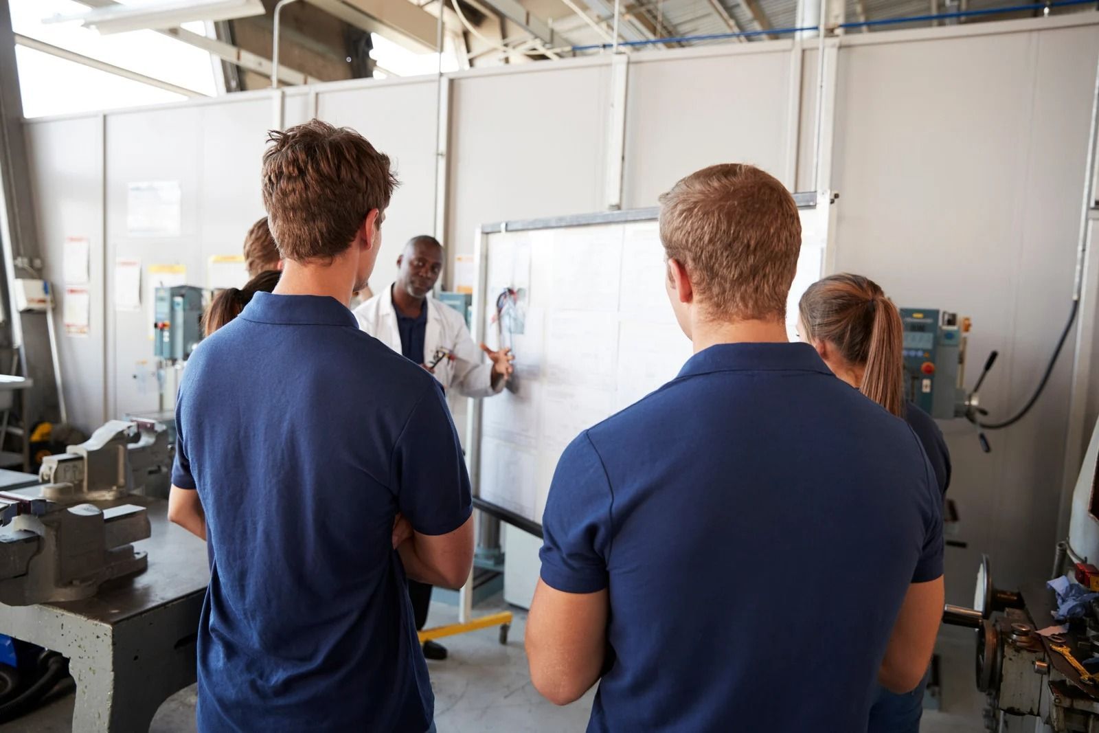 An instructor stands by a whiteboard explaining a concept to students in a vocational workshop with machinery.