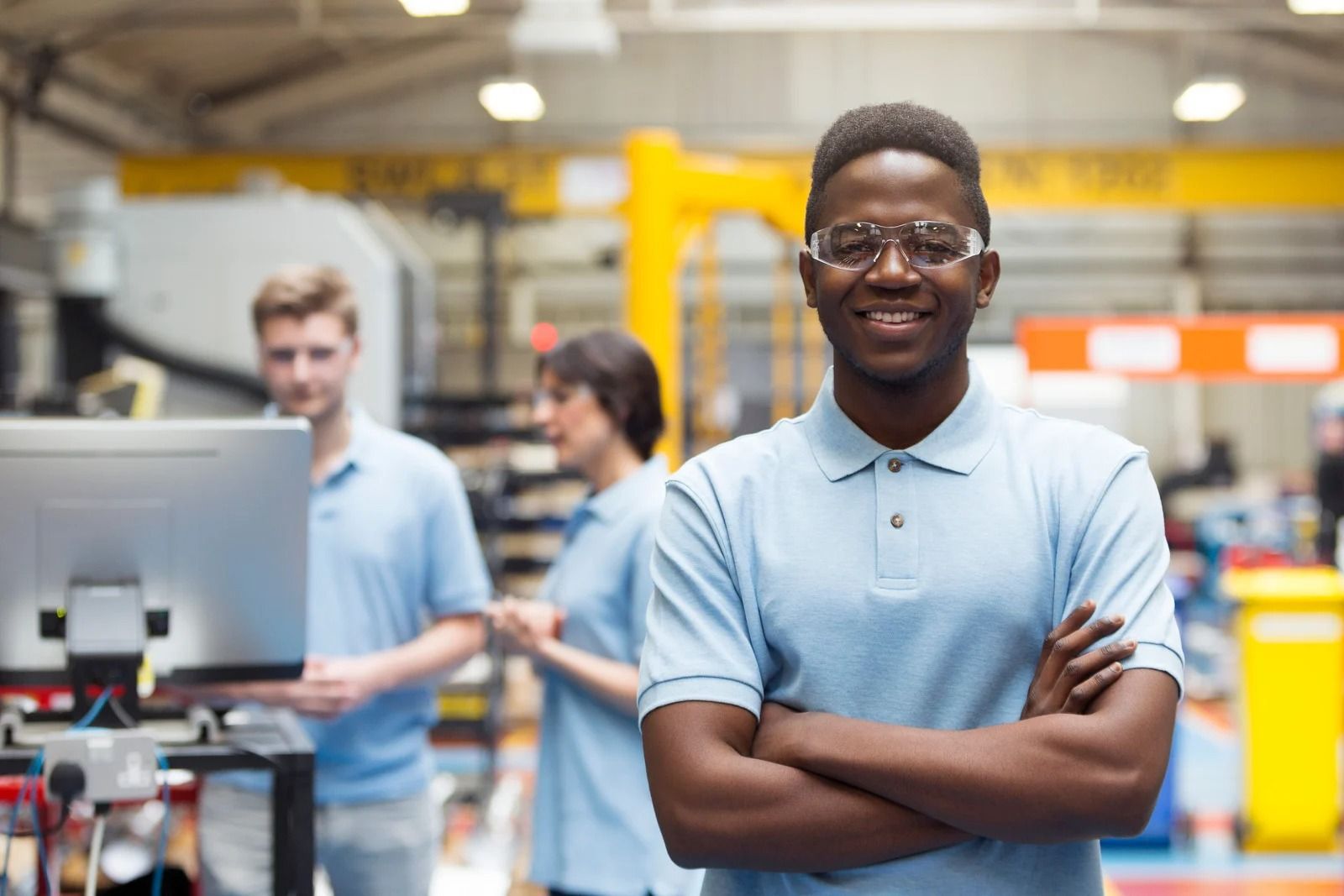 A person in a light blue polo and safety glasses stands with arms crossed in a factory, with colleagues working behind.