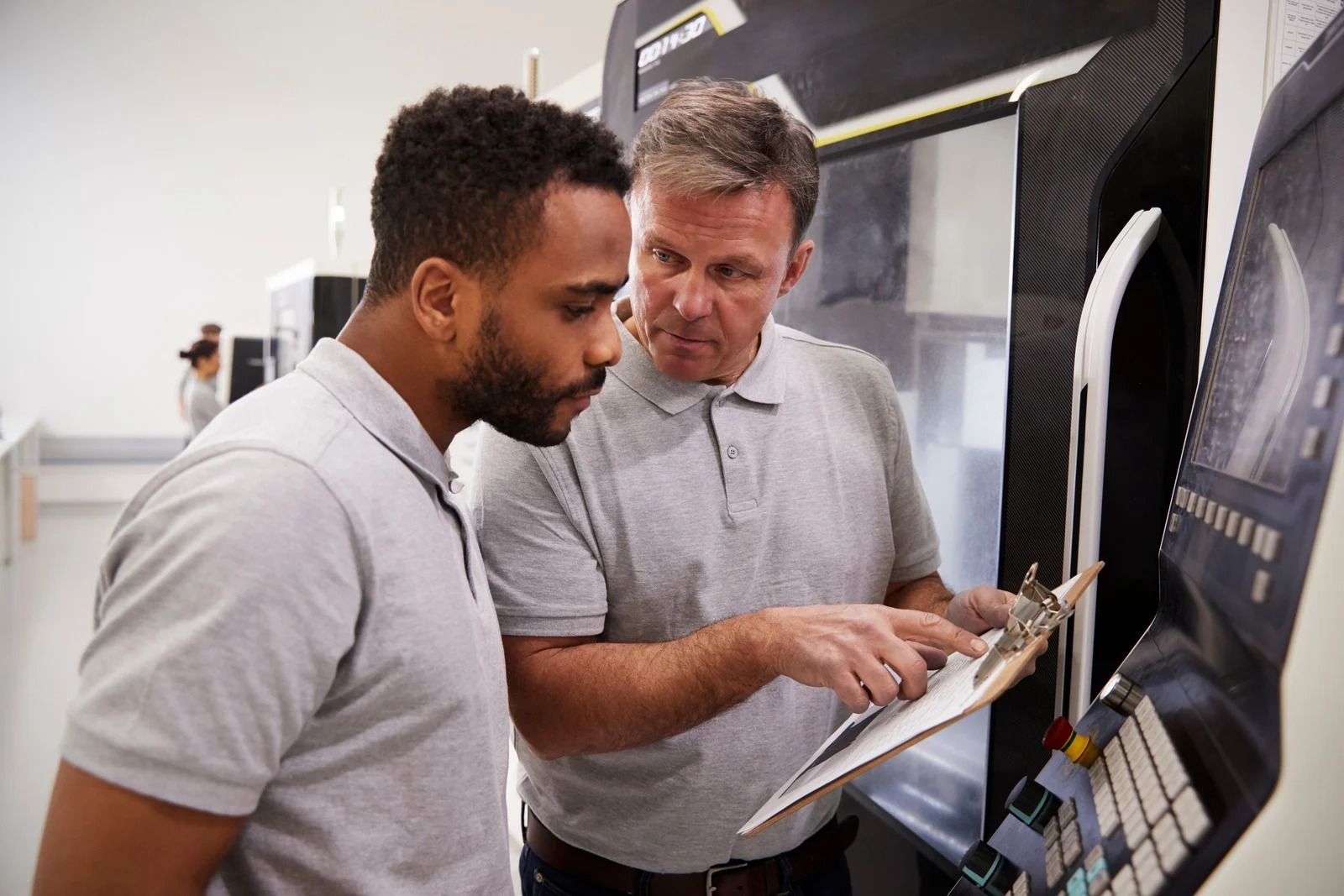 Two people in gray shirts discuss a clipboard near a large industrial machine in a factory setting.