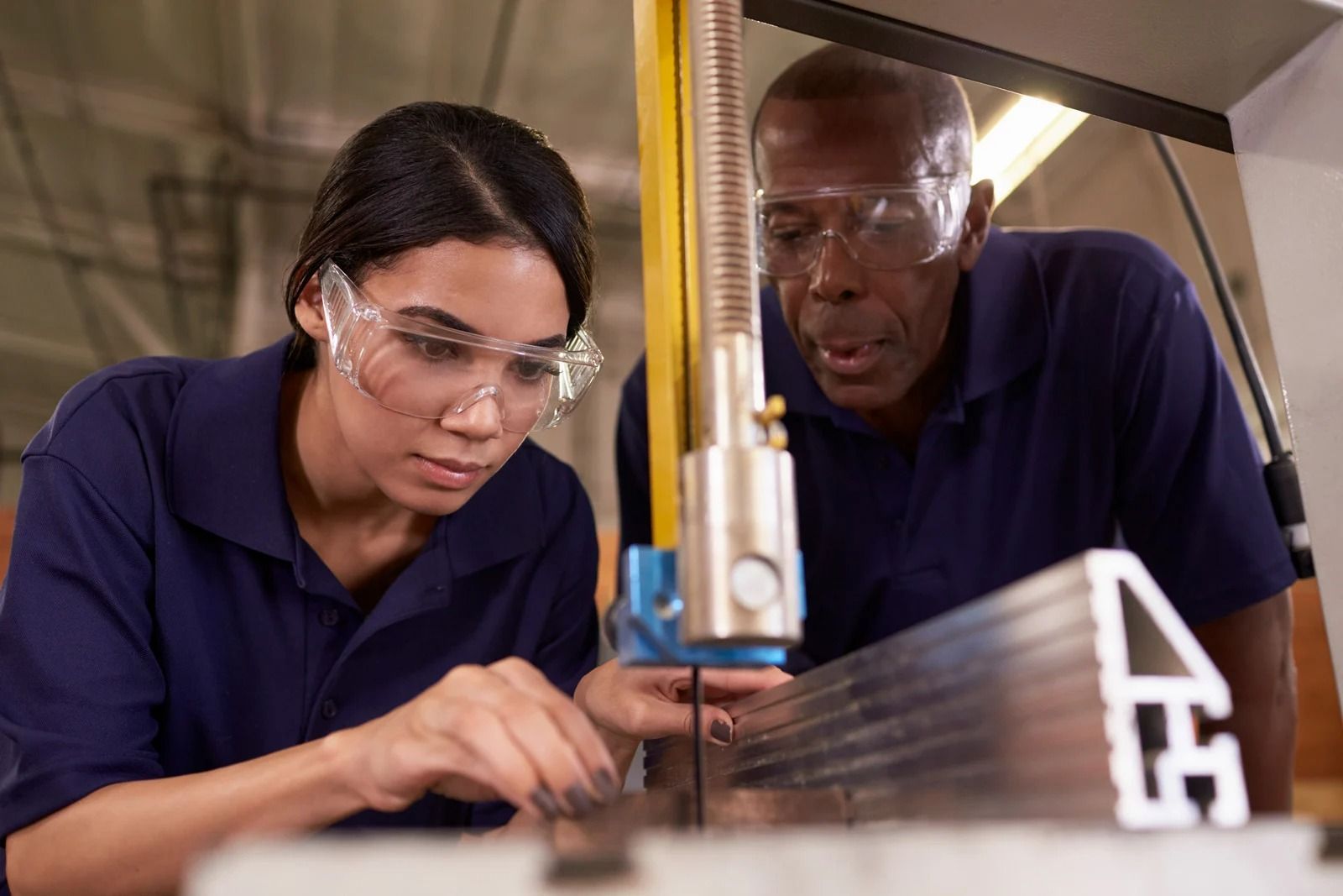 Two people in navy polo shirts and safety glasses carefully operate a metal cutting machine in a workshop.
