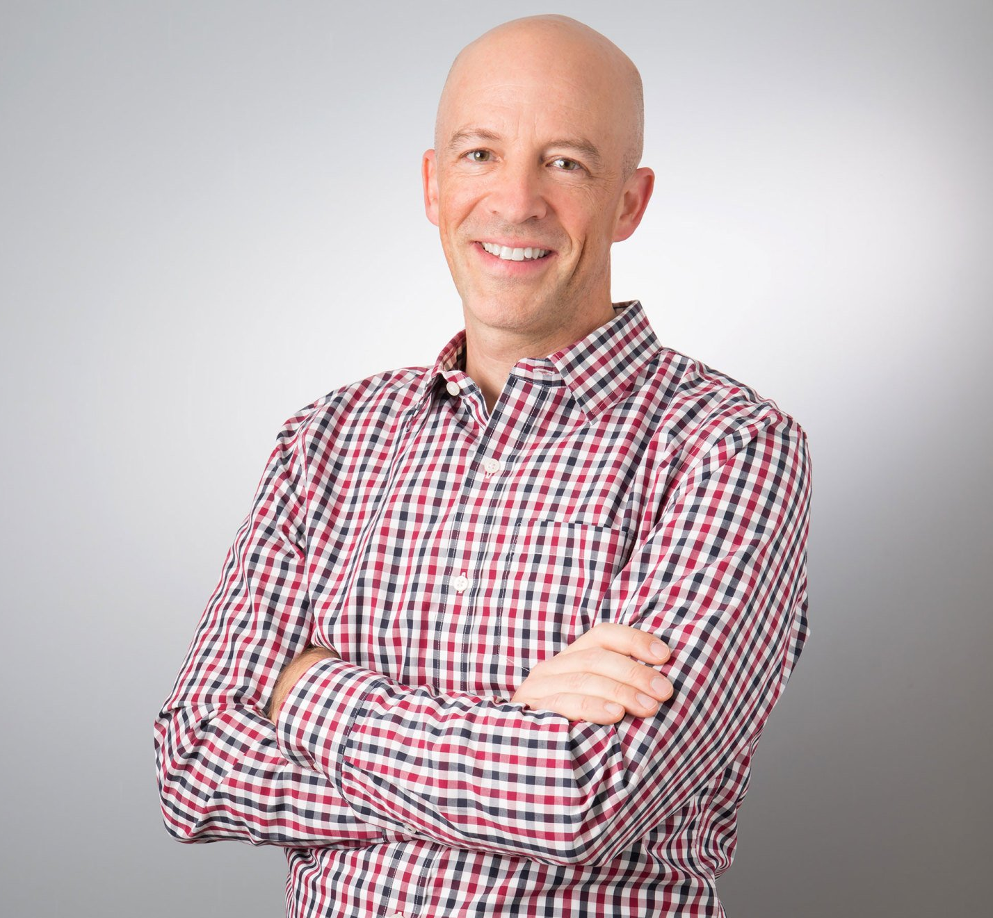 A bald man in a plaid shirt is smiling with his arms crossed.