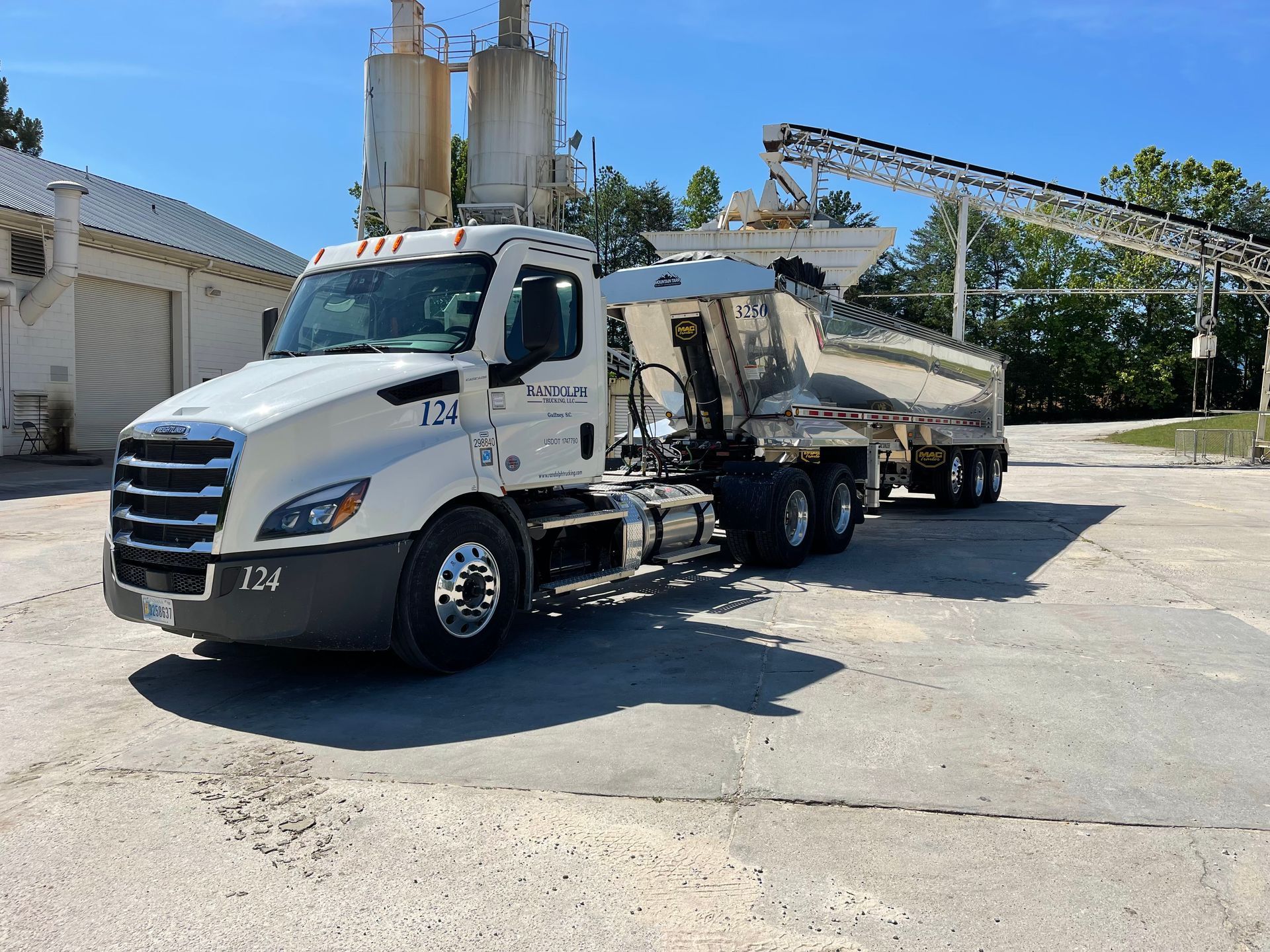 Blue semi-truck with dump trailer, parked on dirt lot near an excavator.