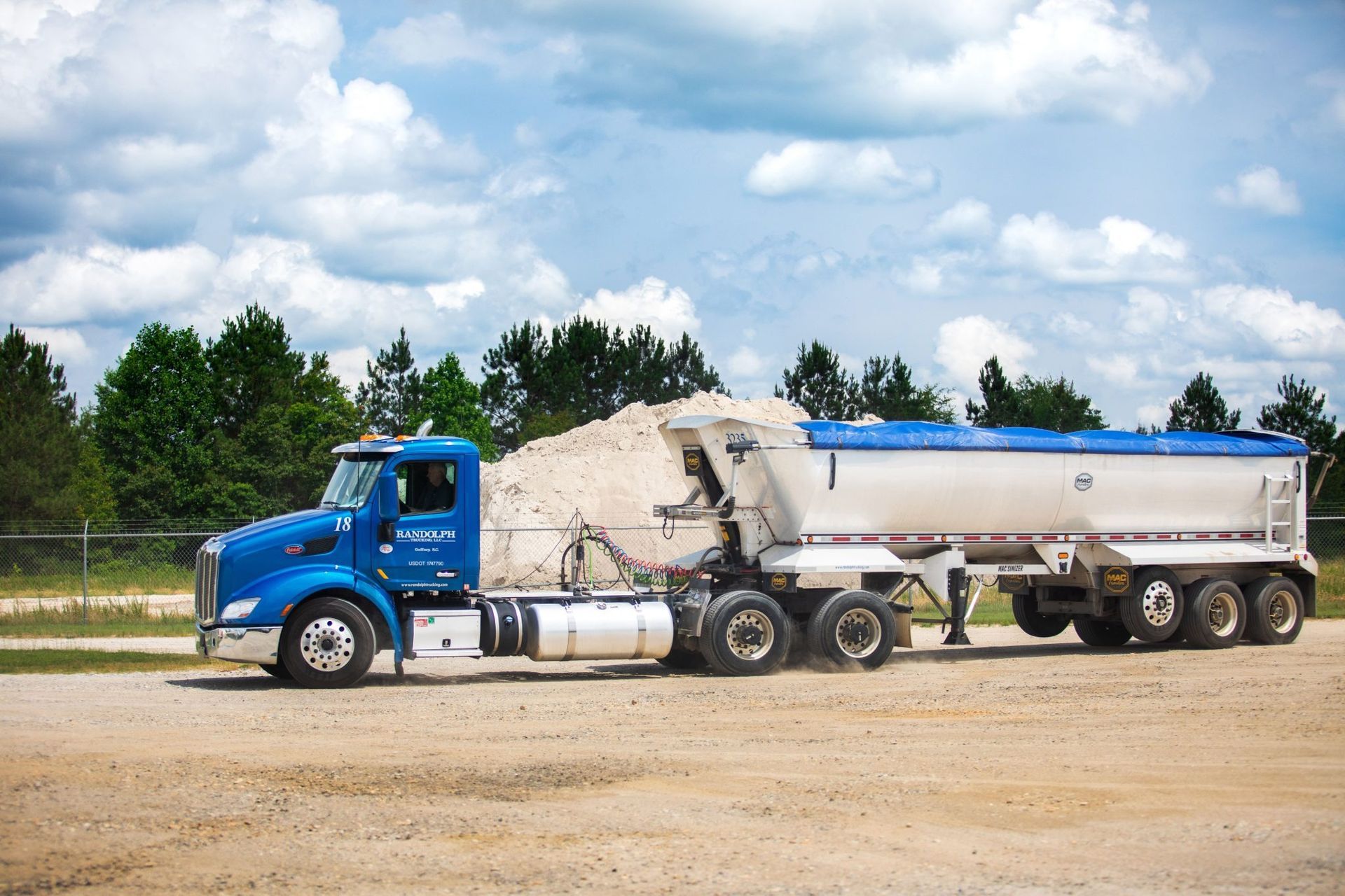 Blue semi-truck with a white trailer, hauling sand. Parked on a dirt road, trees in the background.