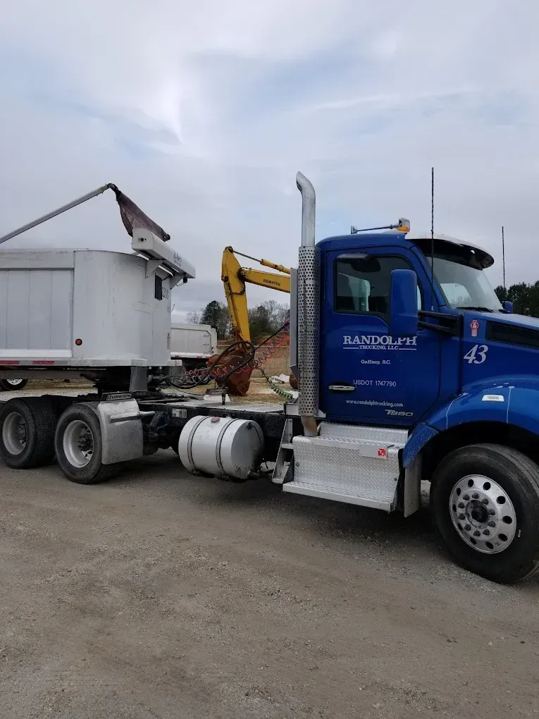 Blue semi-truck with dump trailer, parked on dirt lot near an excavator.
