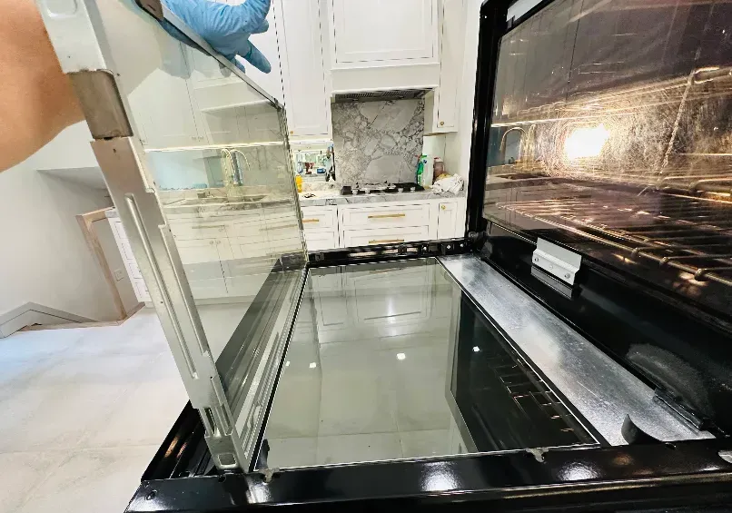 Person removing oven door glass, revealing interior. Kitchen setting with white cabinets and marble backsplash.