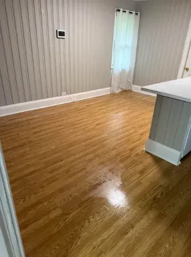 Hardwood floor in room with light gray wainscoting and a white curtained window. A white countertop is visible.