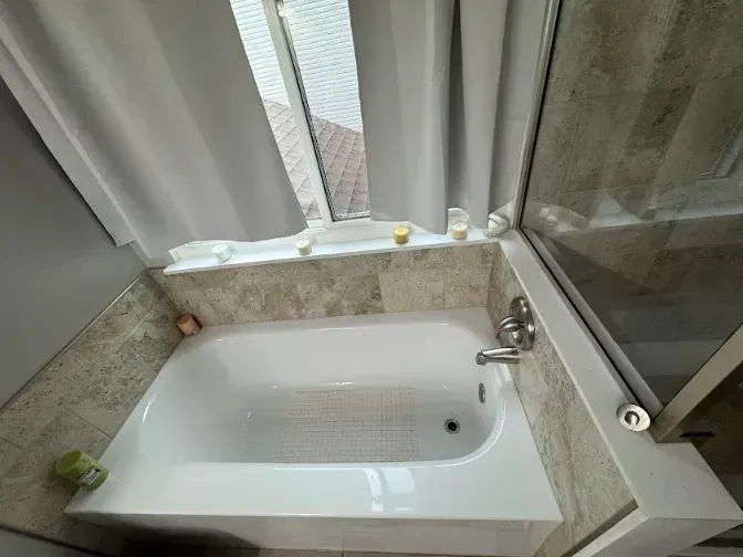 A white bathtub with a window above it, surrounded by beige tiles and a glass shower door.